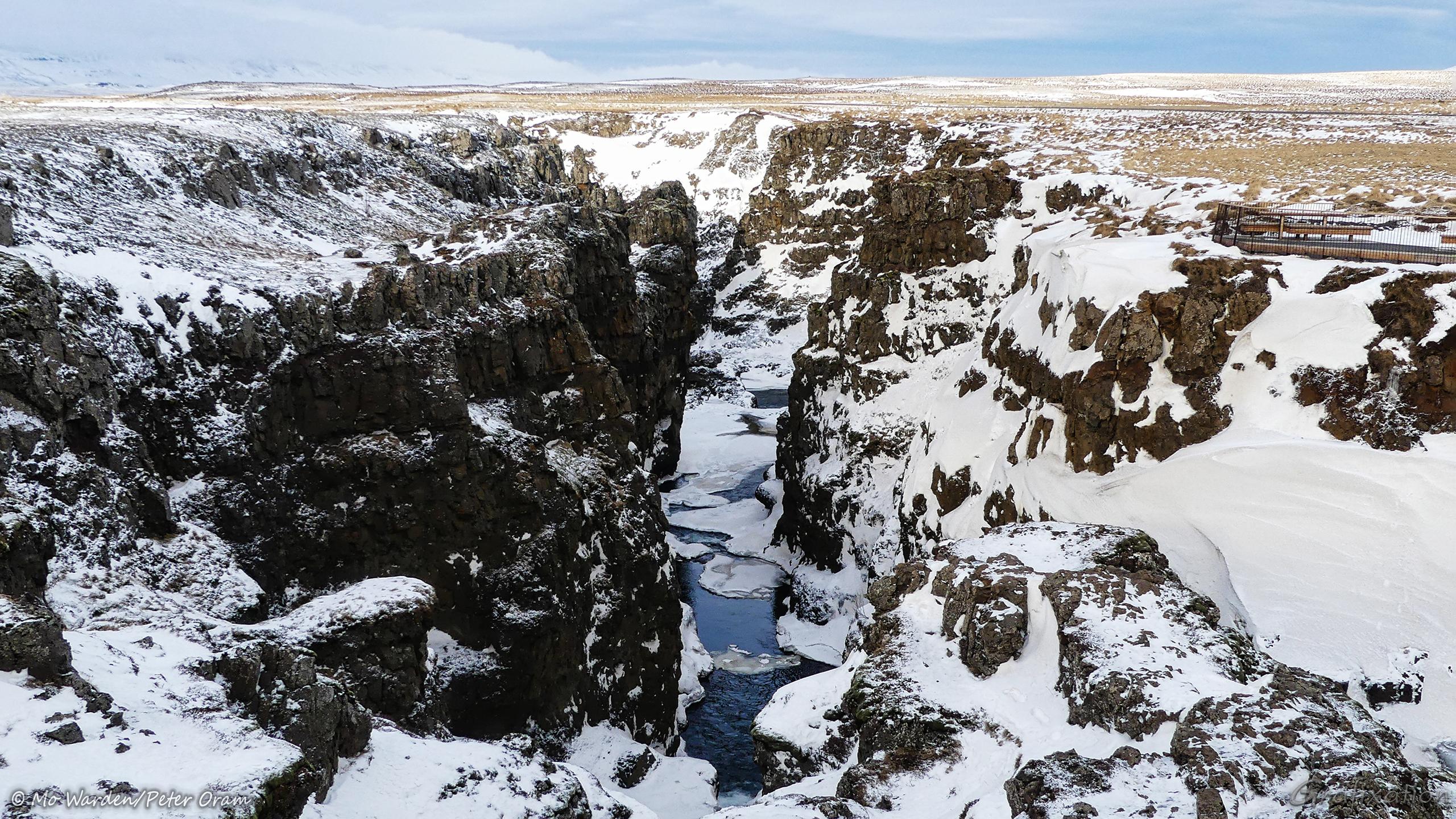 A second colour photo of the canyon looking in the opposite direction. The gorge has dramatically steep walls, quite vertical in places. The small amount of sky that's visible is grey and overcast with snow cloud. On the right is a viewing platform with a barrier and seating which provides some scale. Every ledge that's even close to horizontal is covered with deep snow drifts, but the dark rock of the canyon walls is relatively clear of ice. At the bottom of the gorge, most of the river is frozen over except for small areas where the water surface can be glimpsed.