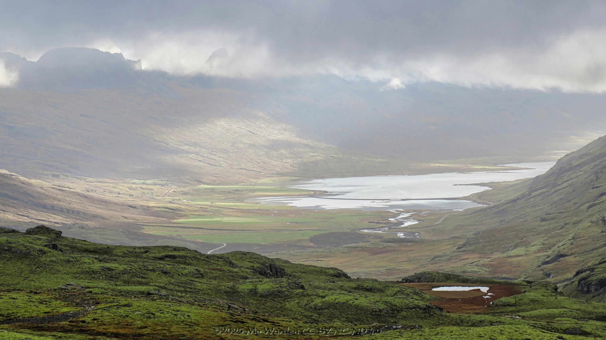 A colour photo of a landscape under a cloudy sky. The viewpoint is from height, overlooking a green valley below leading to the head of a fjord. Some areas are sunlit, showing that the cloud cover isn't complete. The foreground is a wide and rugged mossy ledge with a small tarn on the right. The distant valley below is misty with tonal perspective. The mountains on the opposite side of the valley are mostly obscured by tendrils of high cloud. It's exceptionally lovely.