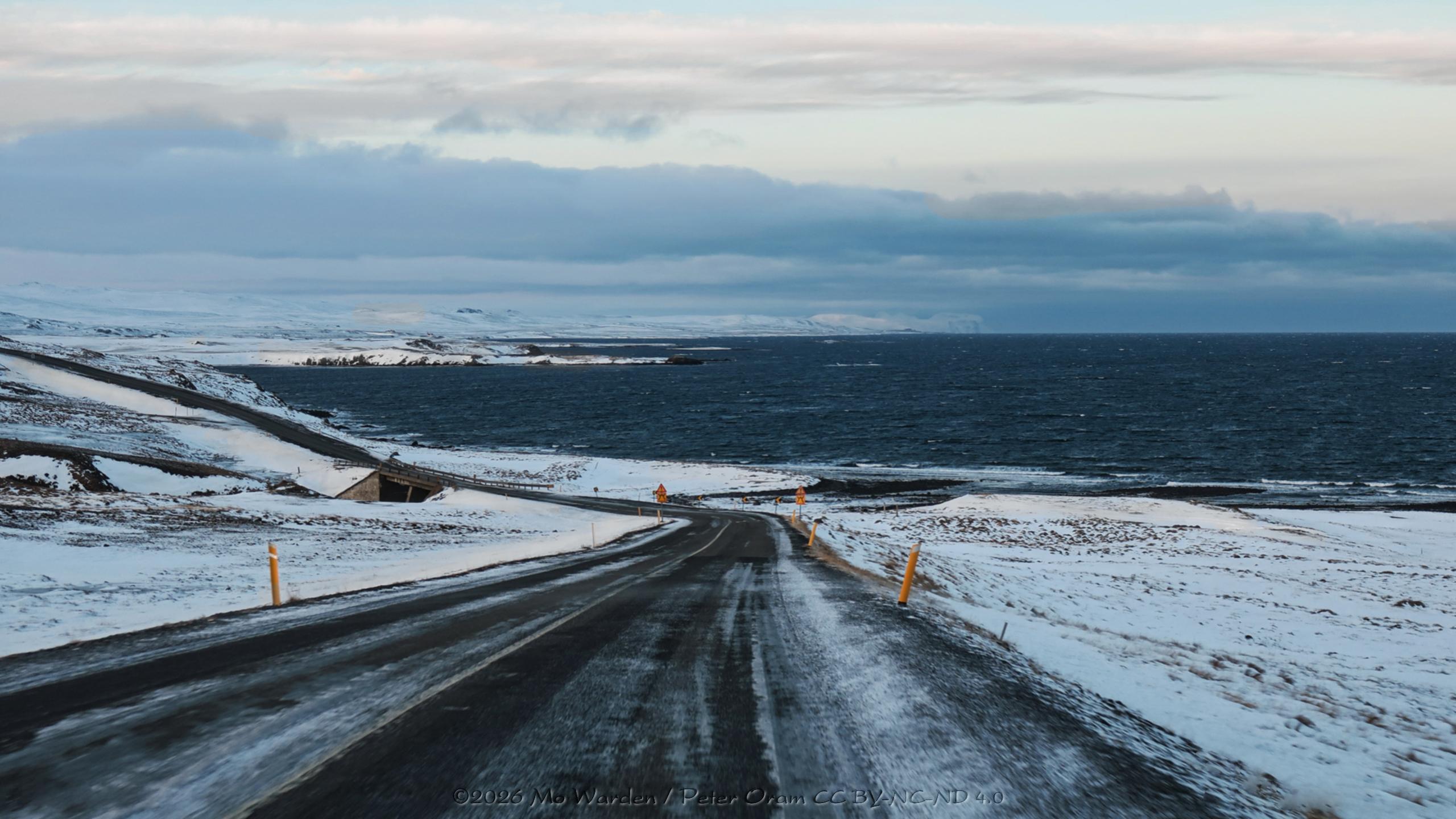 A colour photo of a winter landscape under a mostly cloudy sky. The viewpoint is from a vehicle taking a metalled road down towards the side of a fjord. There are yellow snow poles on either side of the icy black road. Not far ahead the road curves away to the left across a small bridge which has been installed below the road deck. The dark blue waters of the fjord are slightly ruffled and have an edging of white ice as well as small icy patches in the water. The sunlight from the left is very low and is casting a pale pastel pink onto the snow banks facing that direction.