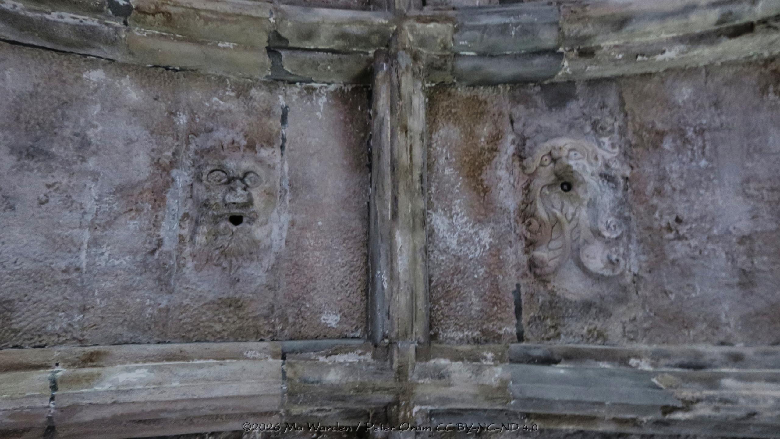 A colour photo of a detail in a vaulted ceiling. On either side of a stone beam are two grotesques, carved in the shape of unhuman faces. The one on the right is of the Green Man, both have wide open eyes. Their mouths are holes from which lamps were hung.