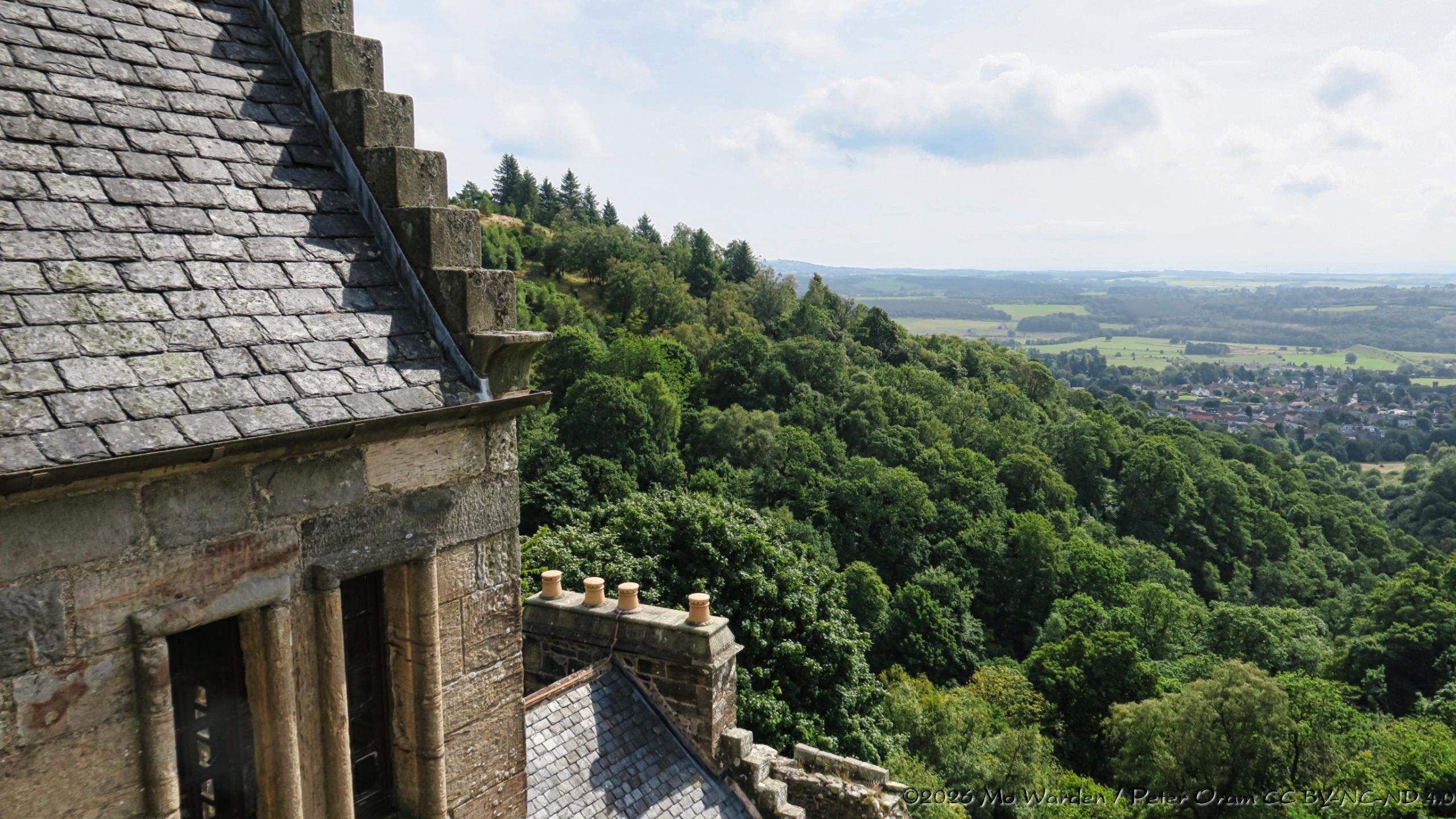 A colour photo of a view from the battlements of the castle. On the left are stone windows, it isn't clear from the shot but they have sash window panes. A slate roof is angled above. Slightly below is another roof with a row of chimney pots, all are closed. The woodland beyond drops dramatically to the valley below.