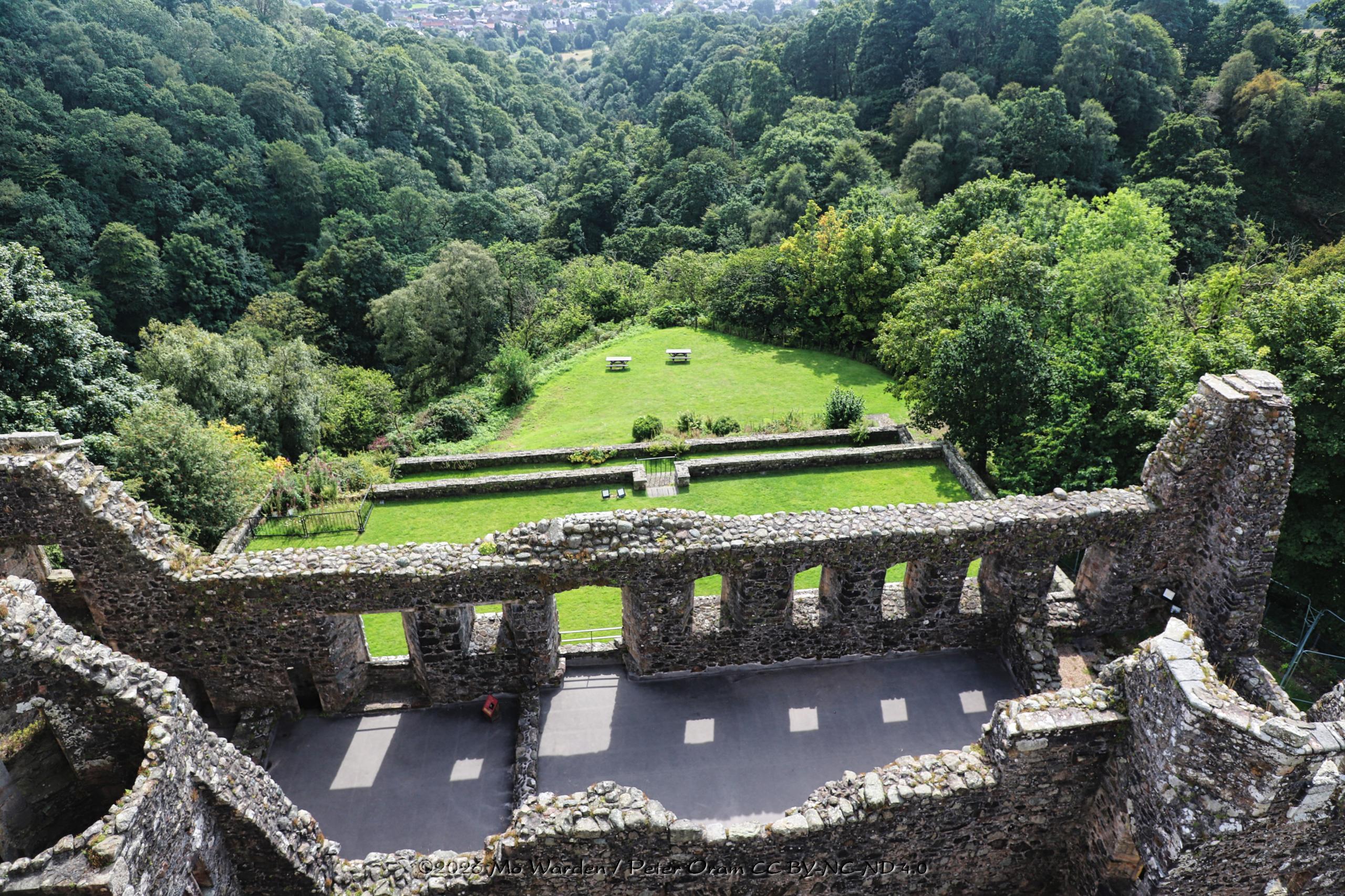 A colour photo of the view from the castle roof looking down into the valley below. Immediately below are grey stone walls, broken at the tops where a roof once covered a cloistered walkway. Beyond that are manicured gardens with embedded ha-ha walls. Below that are thoughtfully placed benches. The ancient woodland stretches away into the distance, lush and verdant.