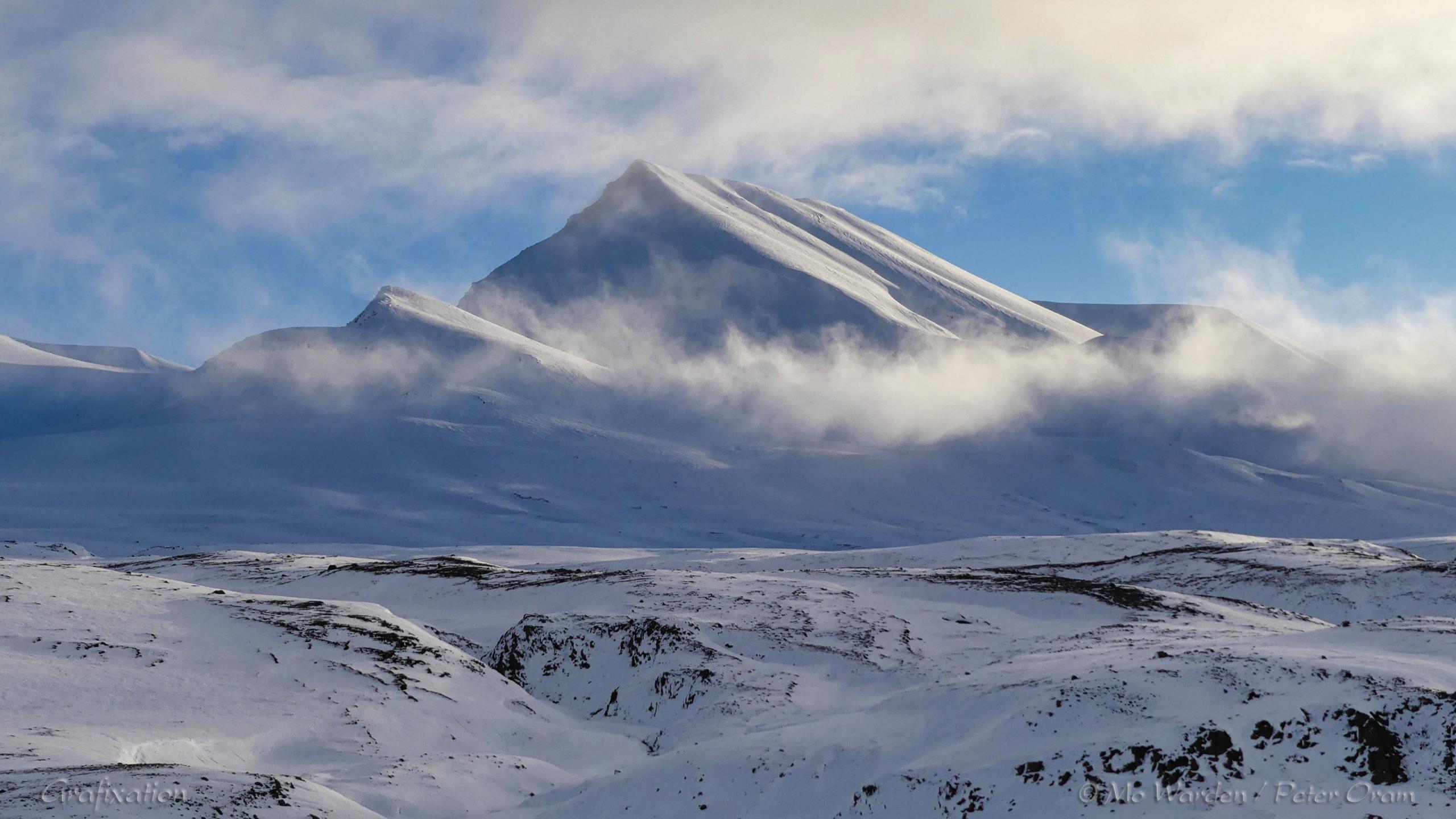 A colour photo of a single mountain pinnacle covered in ice and snow. The foreground is a flat-topped ridge, mostly white with snow but with black rock showing through on ridges where the wind has scoured away the ice. The sky is clear but some low cloud is wrapped around the peak, which is lit from the right. The flanks form a pyramidal shape which is accentuated by strong light on one face.