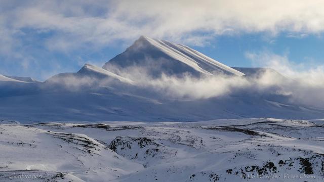 A colour photo of a single mountain pinnacle covered in ice and snow. The foreground is a flat-topped ridge, mostly white with snow but with black rock showing through on ridges where the wind has scoured away the ice. The sky is clear but some low cloud is wrapped around the peak, which is lit from the right. The flanks form a pyramidal shape which is accentuated by strong light on one face.
