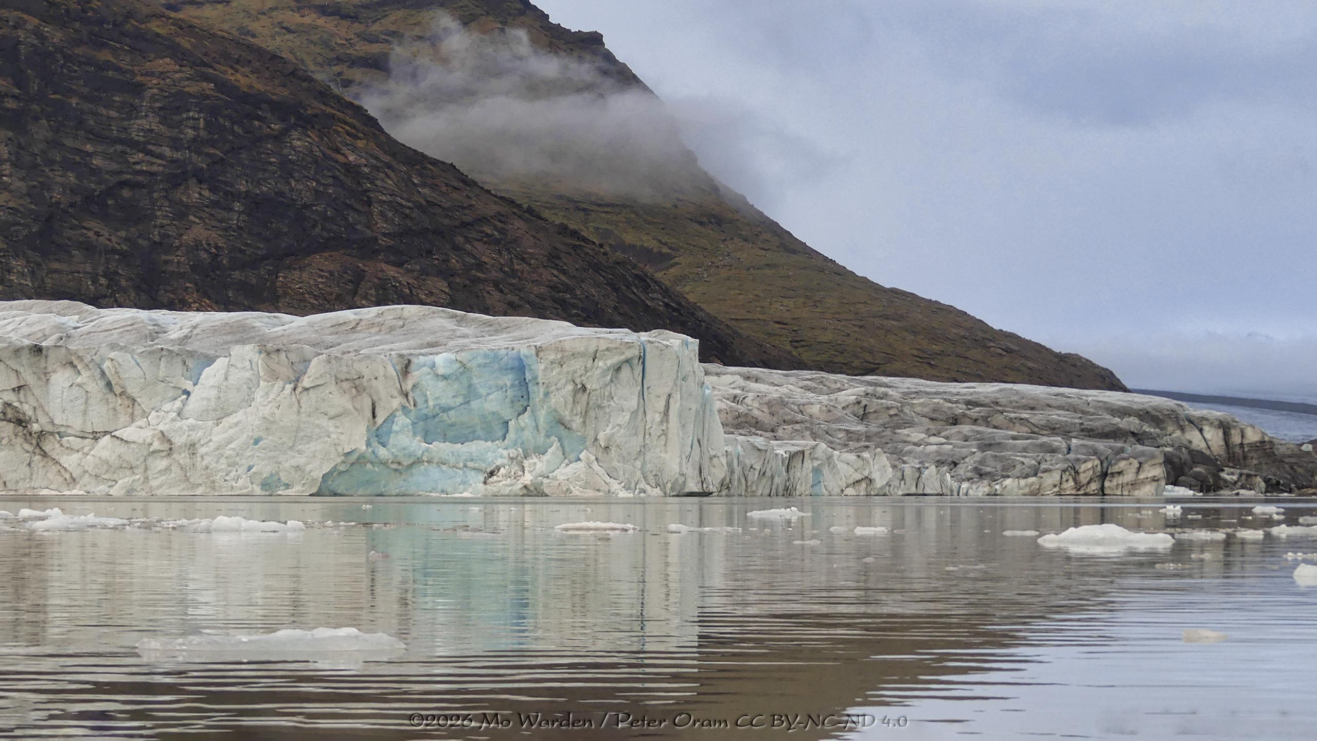 A colour photo of a rippled lake with ice floating at its surface. In the background are mountain flanks, between them are wisps of thin cloud. The sky is overcast but not heavily. In the centre of the shot is a line of turquoise ice blocks forming the calving face of a glacier. In many places the ice is brown or black from volcanic deposits, but deep within the ice is a lot of cyan colouring. The mountains and ice front are reflected in the water.