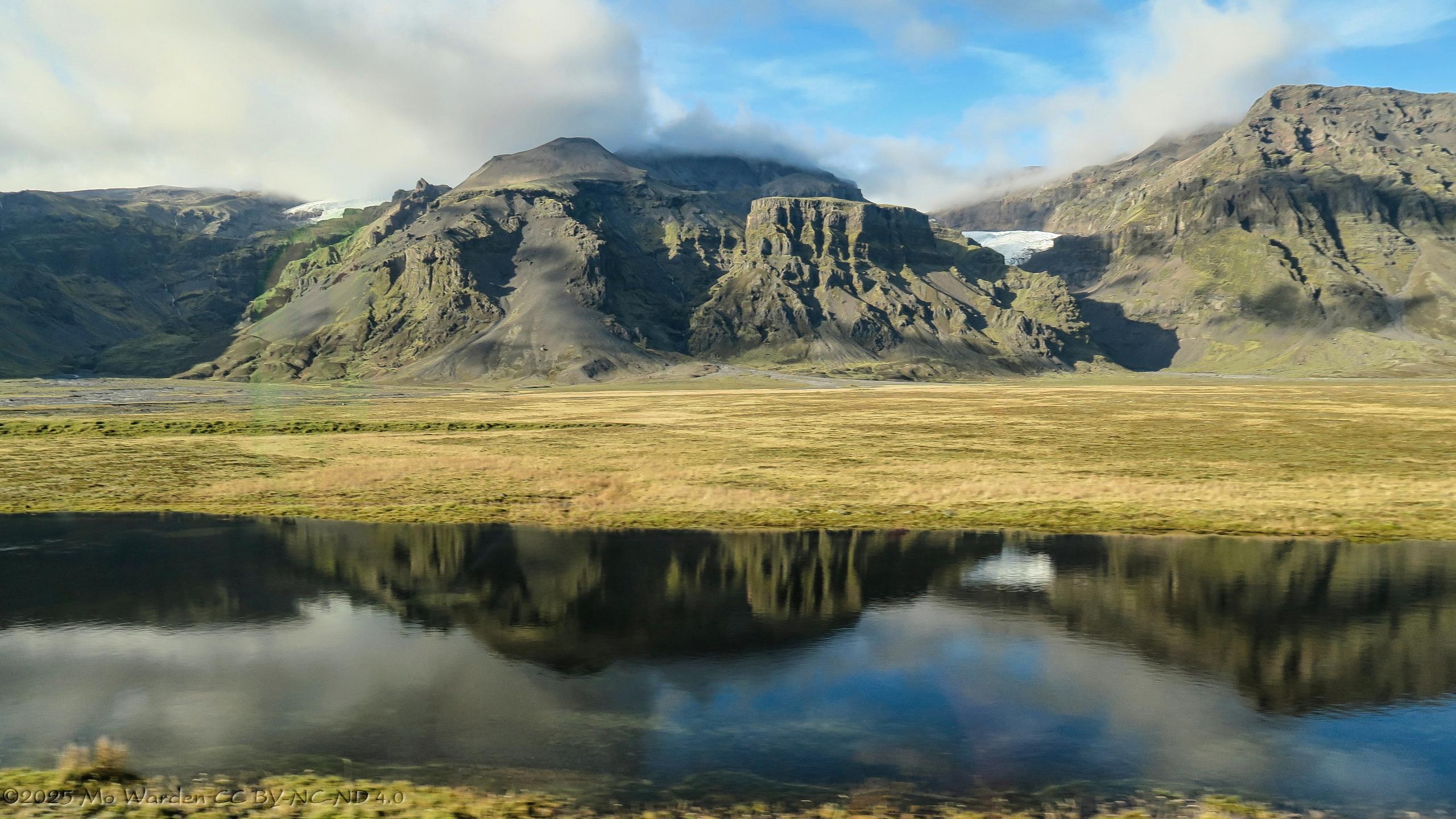 A colour photo of mountains with glaciers dropping between them. The sky is cloudy but with clear patches and the sunlight is from the left and behind the viewer. Craggy greyish brown rocks stand between the tongues, at their foot is a flat grassy area. In the foreground is a body of shallow water, probably flooding, which is reflecting the scene.