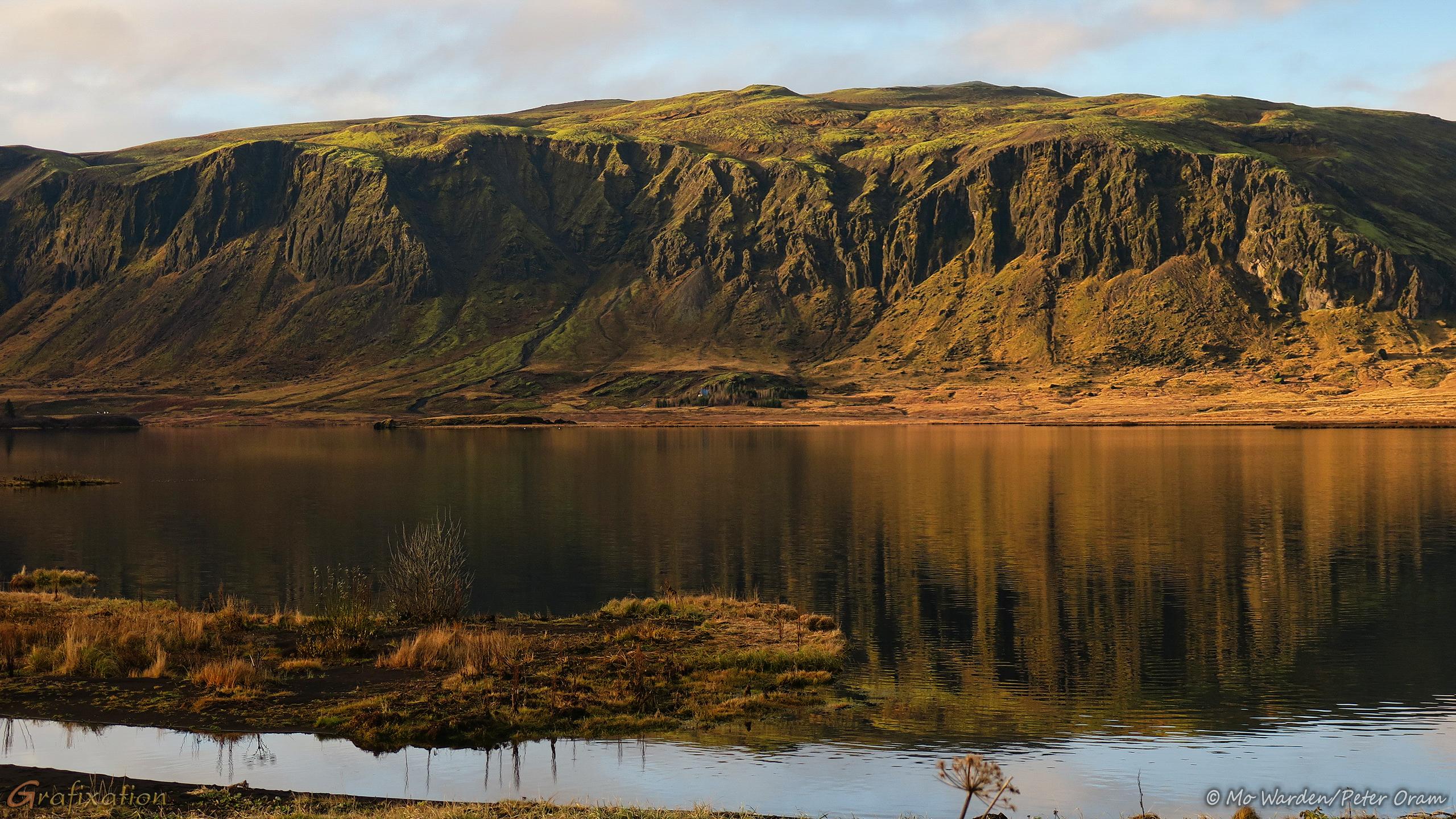 A colour photo of an almost vertical mountainside. Soft warm light from the left is warming the colour of the rocks and picking out detail as contrasts and shadows. The sky is lightly clouded. The foreground is a body of water with a marshy edge of black sand with scrub vegetation. The water surface is softly rippled by a breeze, and is reflecting a distorted image of the mountainside.