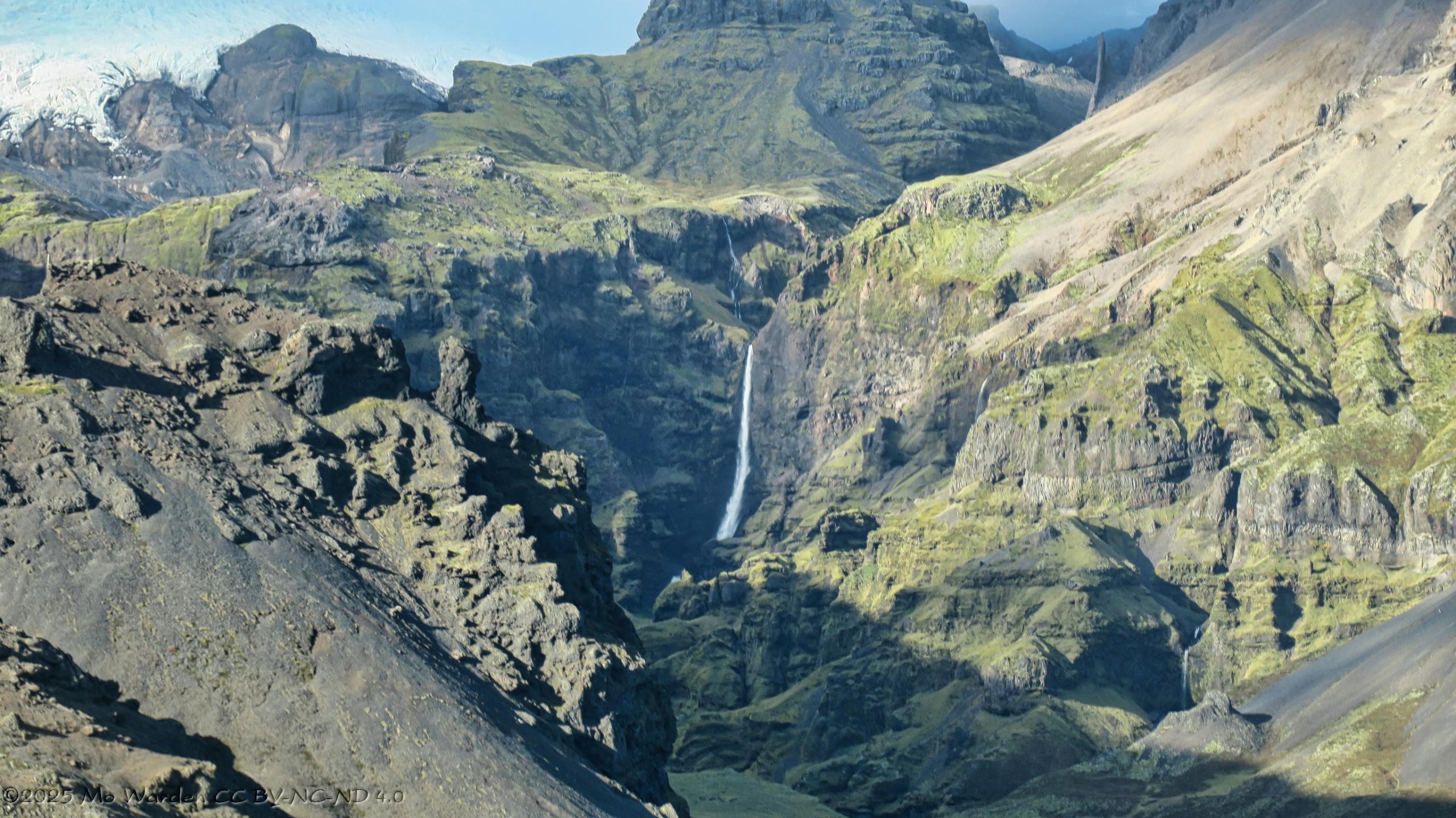 A colour photo of a mountain cut by a deep valley. The mountainside continues up to the top of the shot, no sky is visible. The bright sunlight is from the left. At the top left of the photo is the snout of a glacier, high in the mountains. An unseen river is flowing through the dramatic gulley, reaching a shelf in the rock and dropping as a lovely horsetail waterfall. Just before it disappears from view behind the rocks, it splashes against the rock face and flows down it.