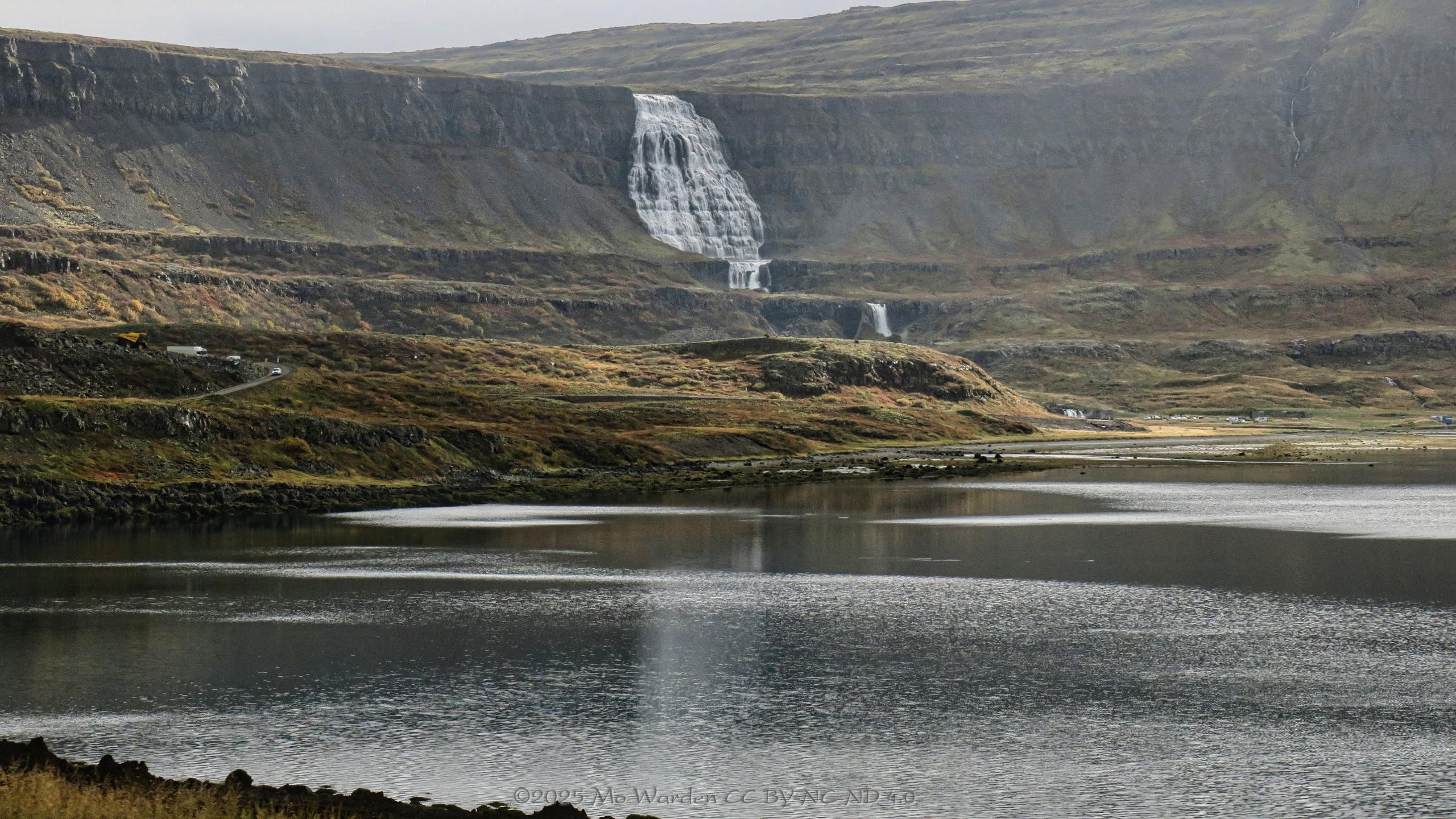 A colour photo of an autumnal landscape. The foreground is a large body of rippled water, leading to uneven banks on the other side. Traffic on the road at the left and parked on the right are lending scale. Around the head of the fjord is an almost vertical bluff of grey rock with mossy patches. Sunlight from the right is striking parts of it. Near the centre is a spectacular white fan waterfall, dropping from the top of the cliff in steps down to the fjord.