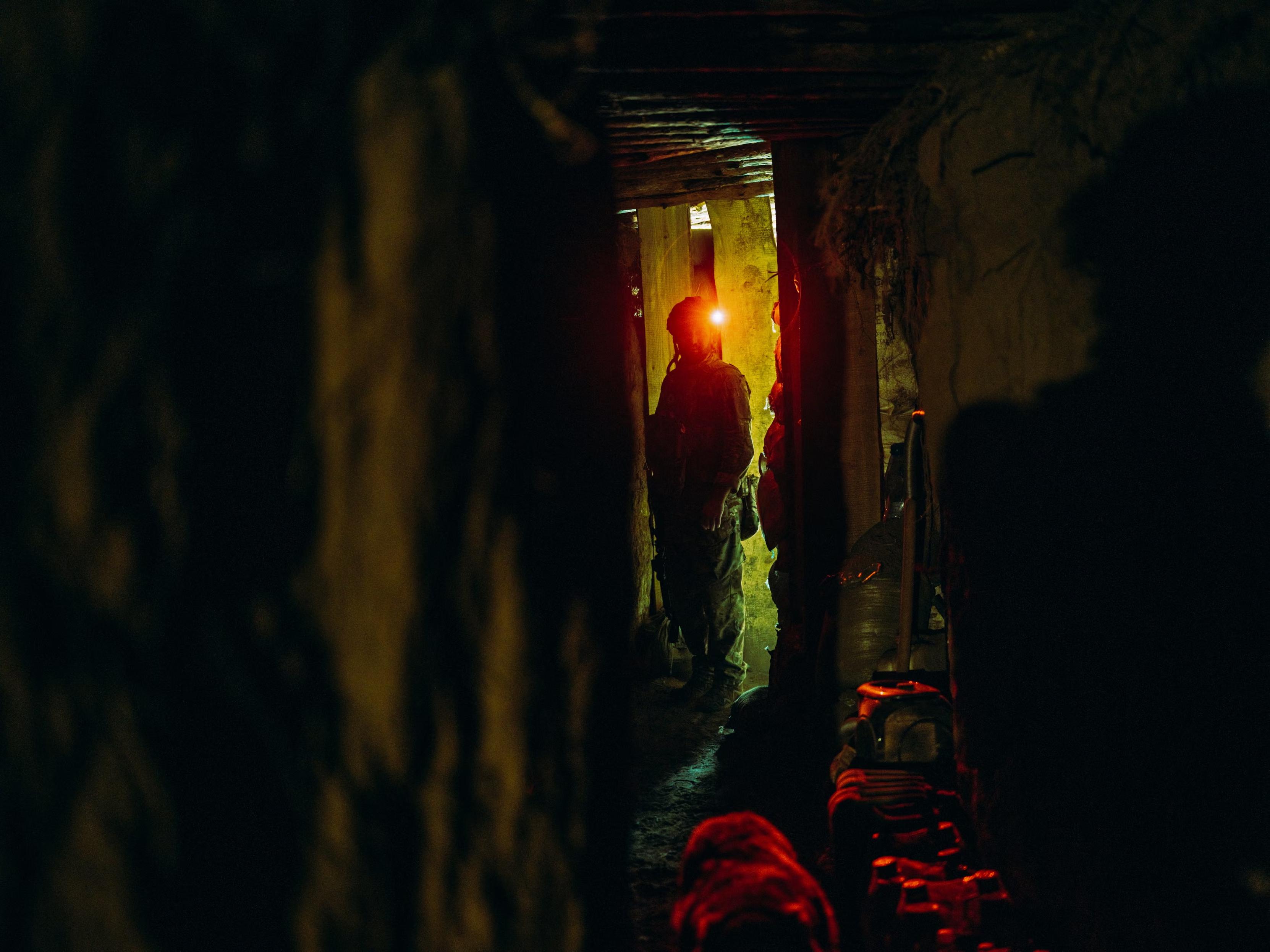 A soldier watches towards the camera with his helmet light on in a dark corridor in the dugout. The Guardian caption: ’ Dzhango, a shooter, gets ready before the daily shift ’