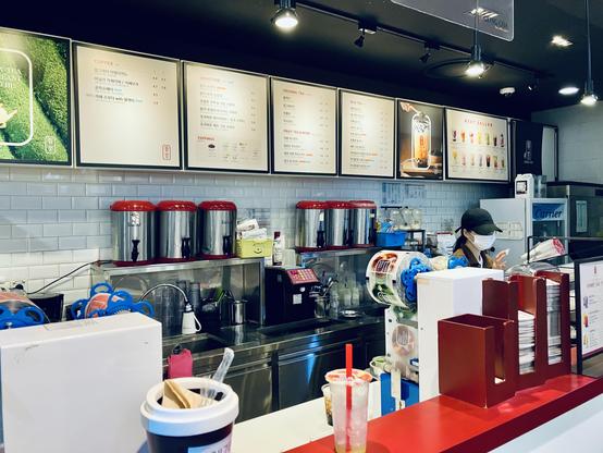 A modern bubble tea shop interior with a clean, minimalist design. Illuminated menu boards line the back wall, displaying coffee, smoothies, original teas, milk teas, fruit teas, toppings, and best-selling drinks. Beneath the menus, several large stainless steel tea urns with red lids sit on the preparation counter alongside stacks of plastic cups, syrup bottles, sealing film rolls, and drink-making equipment. A masked employee wearing a black cap works behind the counter. In the foreground, a red service counter holds napkin dispensers, a takeaway coffee cup, and a partially finished iced drink with a red straw. Bright spotlights illuminate the space, while white subway tile walls and black ceilings create a modern café aesthetic.