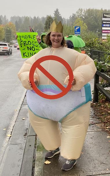 A woman wearing a crown and an inflatable diaper baby costume, without the head, carrying a very large circular 
