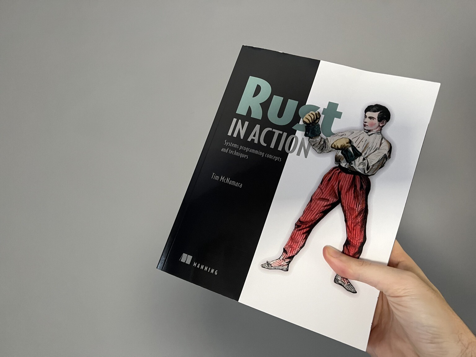 A copy of the book Rust in Action being held up against a grey wall. The cover of the book depicts the sketch on a man in red trousers and a white shirt. His fists are raised.
