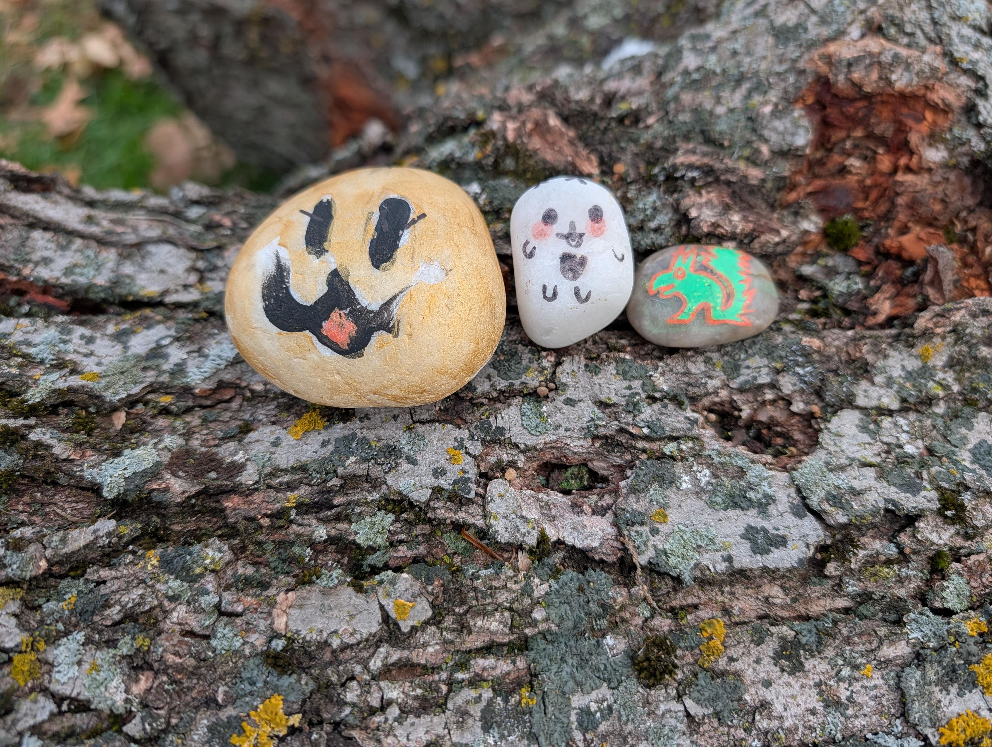 Three painted rocks with faces sit on a tree trunk.