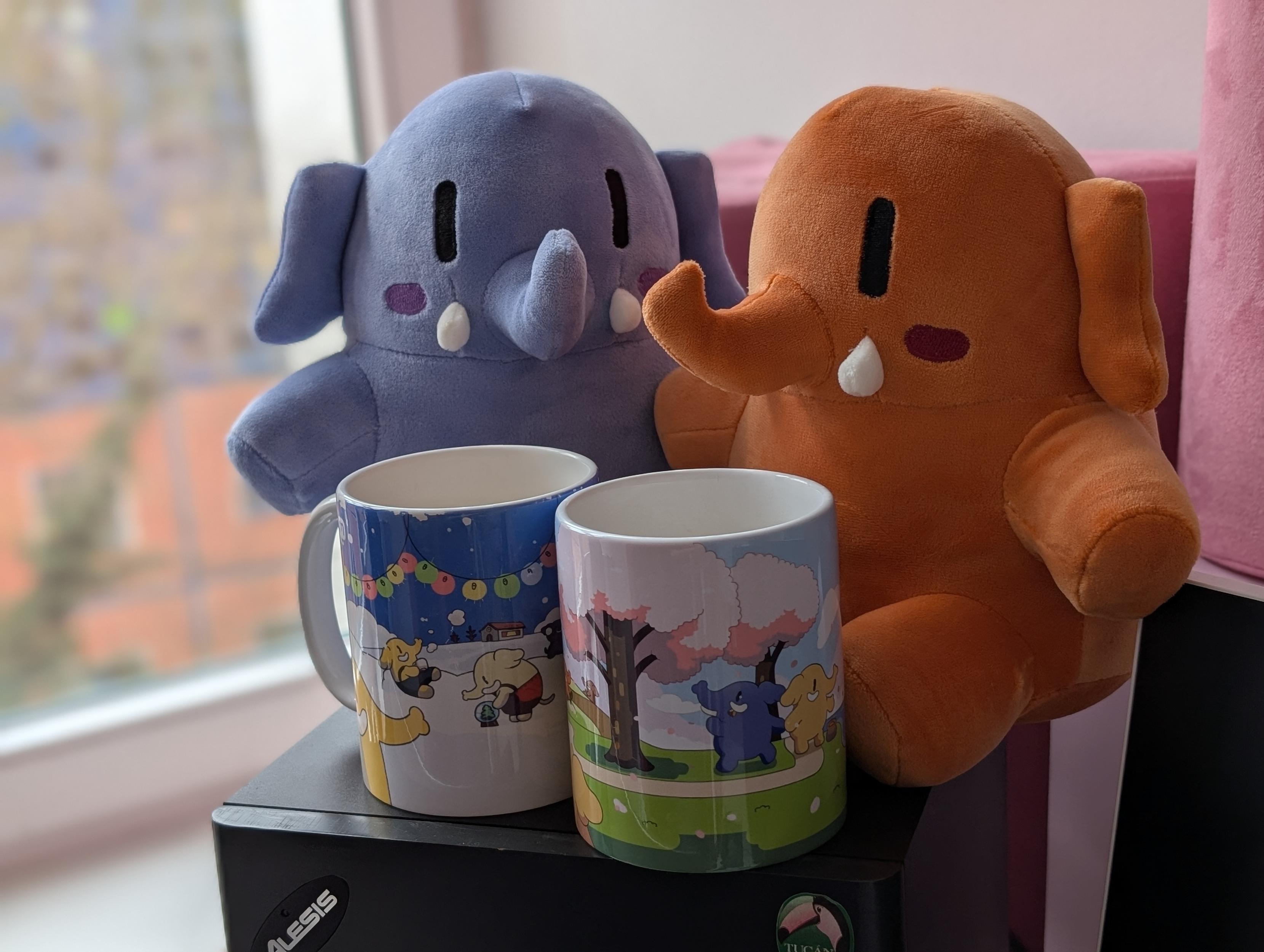 Two Mastodon plushies, one blue, one orange, sitting behind Mastodon-branded coffee mugs. One mug has a dark blue snow & winter print, the other a pink & green spring print.