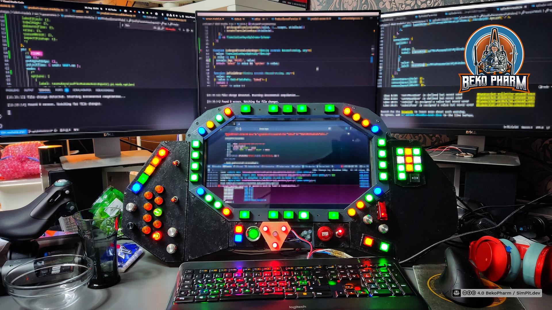 A complex, multi-monitor setup with a custom-built control panel in the foreground. Three large computer monitors are positioned side-by-side. The custom control panel is a prominent, dark-colored, polygonal device with numerous illuminated buttons and switches arranged around a central screen. A flightstick controller is partially visible but not in use currently.
