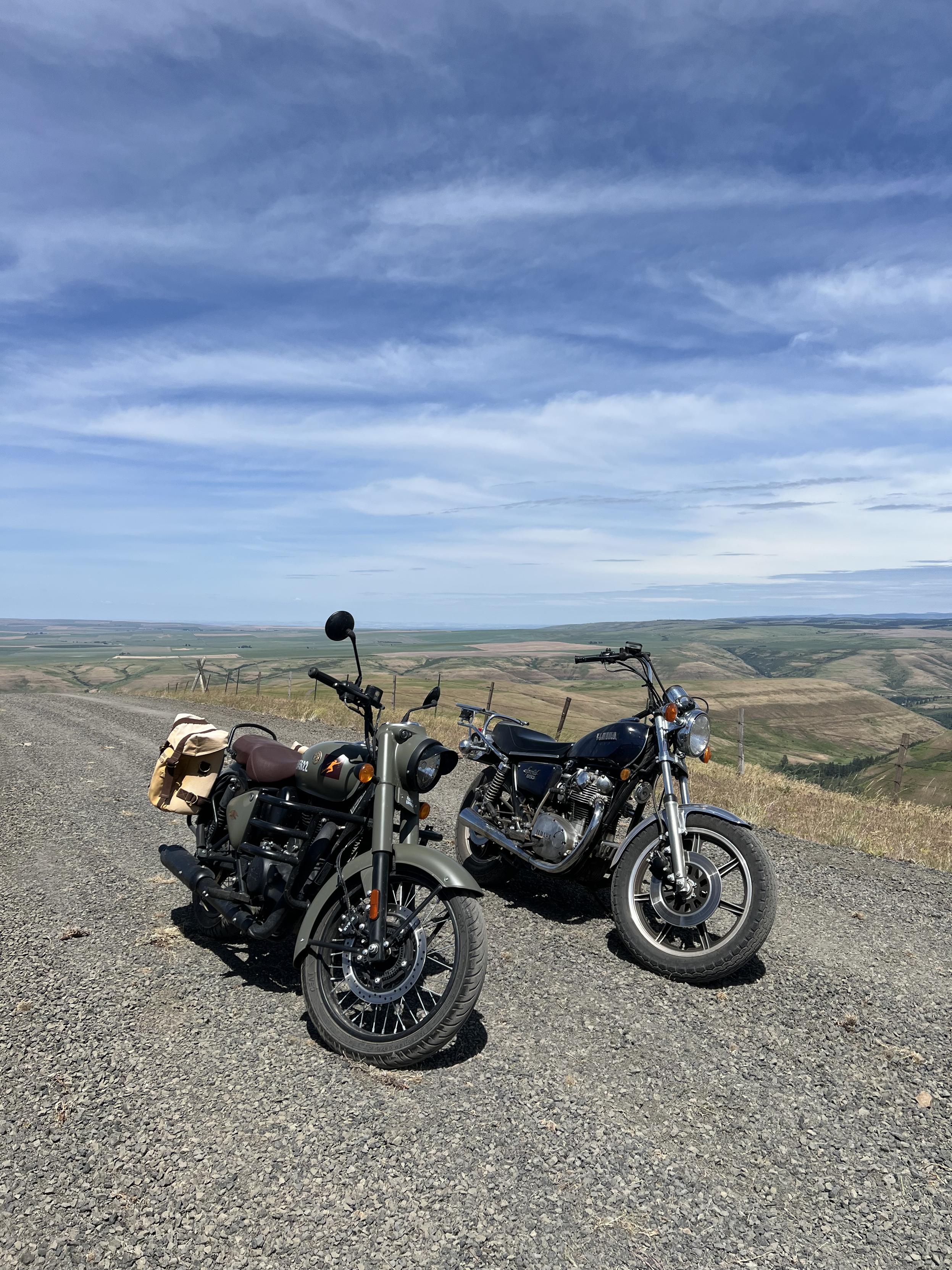 Two motorcycles sitting on a gravel road. The closer left most bike is green, the further bike is blue. Rolling hills are visible in the distance.