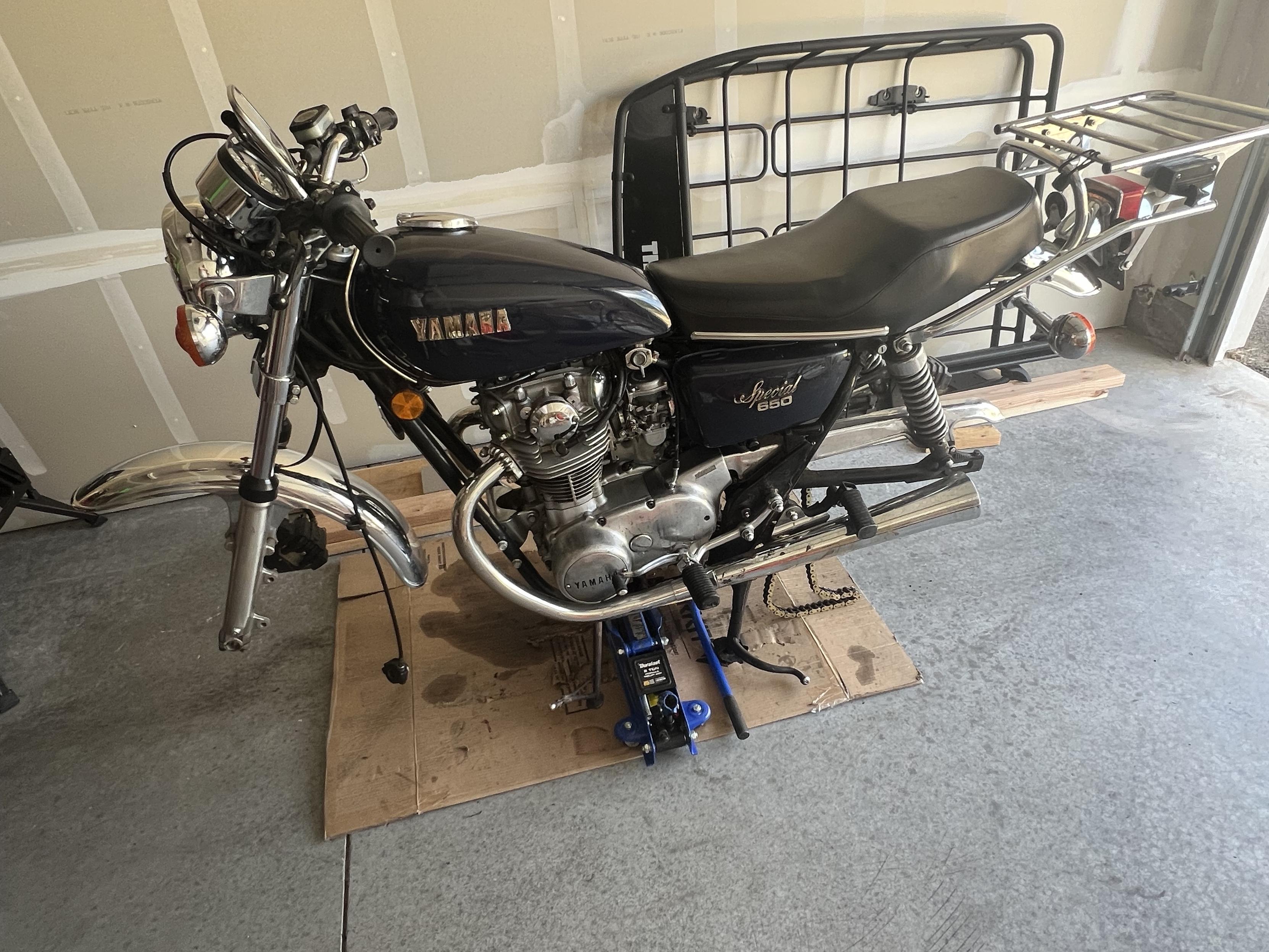 A blue Yamaha motorcycle with its wheels removed. It’s balancing on the center stand and a jack stand. Beneath the bike is a large piece of oil stained cardboard.