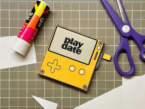 craft Playdate console on a cutting mat, next to a glue stick, scissors, and some cut paper