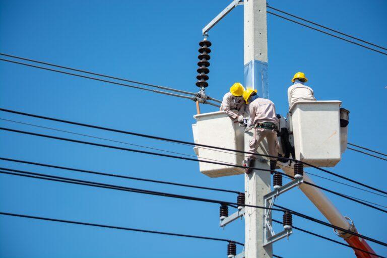 Three people with yellow hard hats work high in the air on what appear to be high voltage power lines.