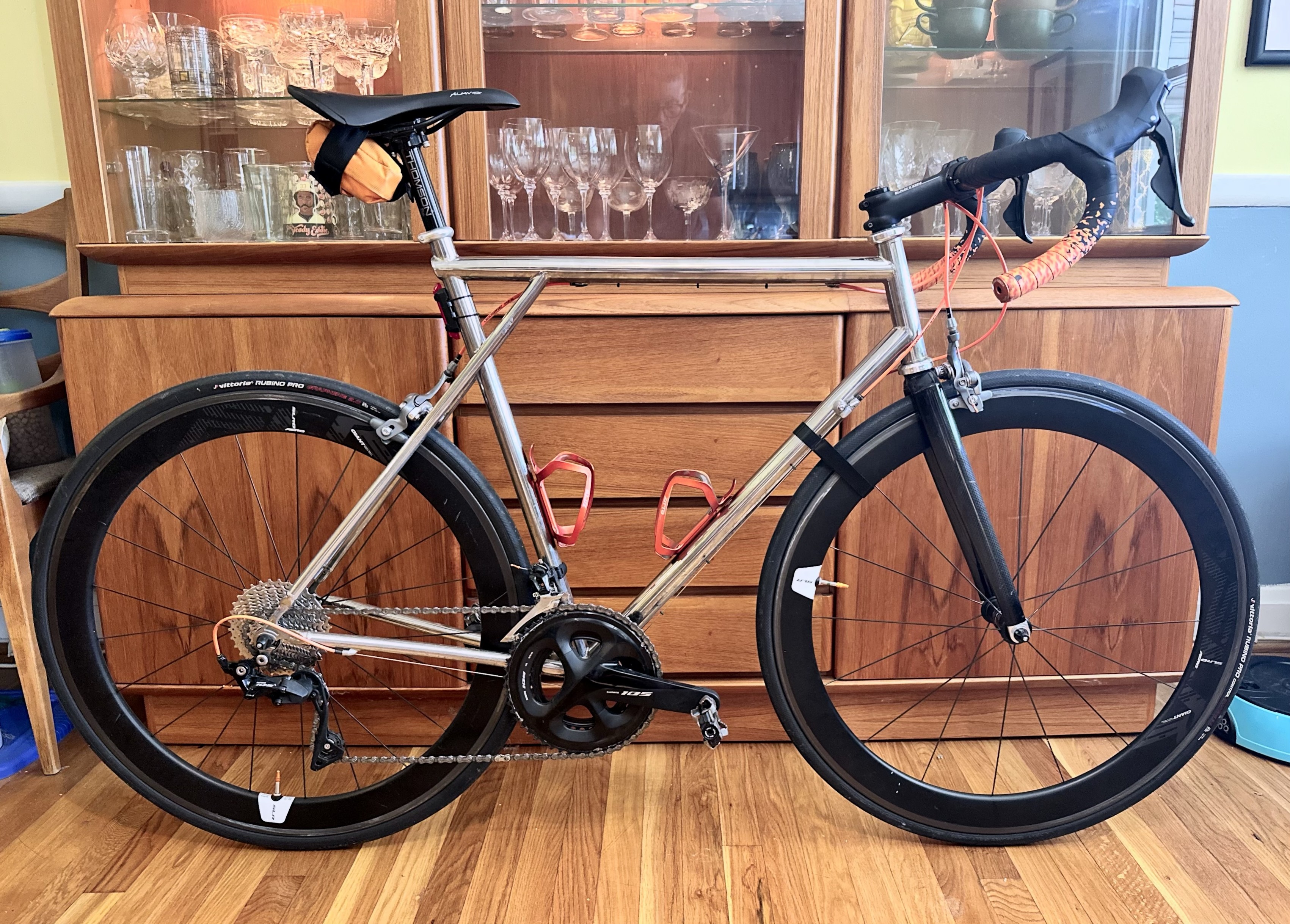 A polished titanium bike frame from 1995 with black components and orange accents in front of a mid-century china cabinet. 