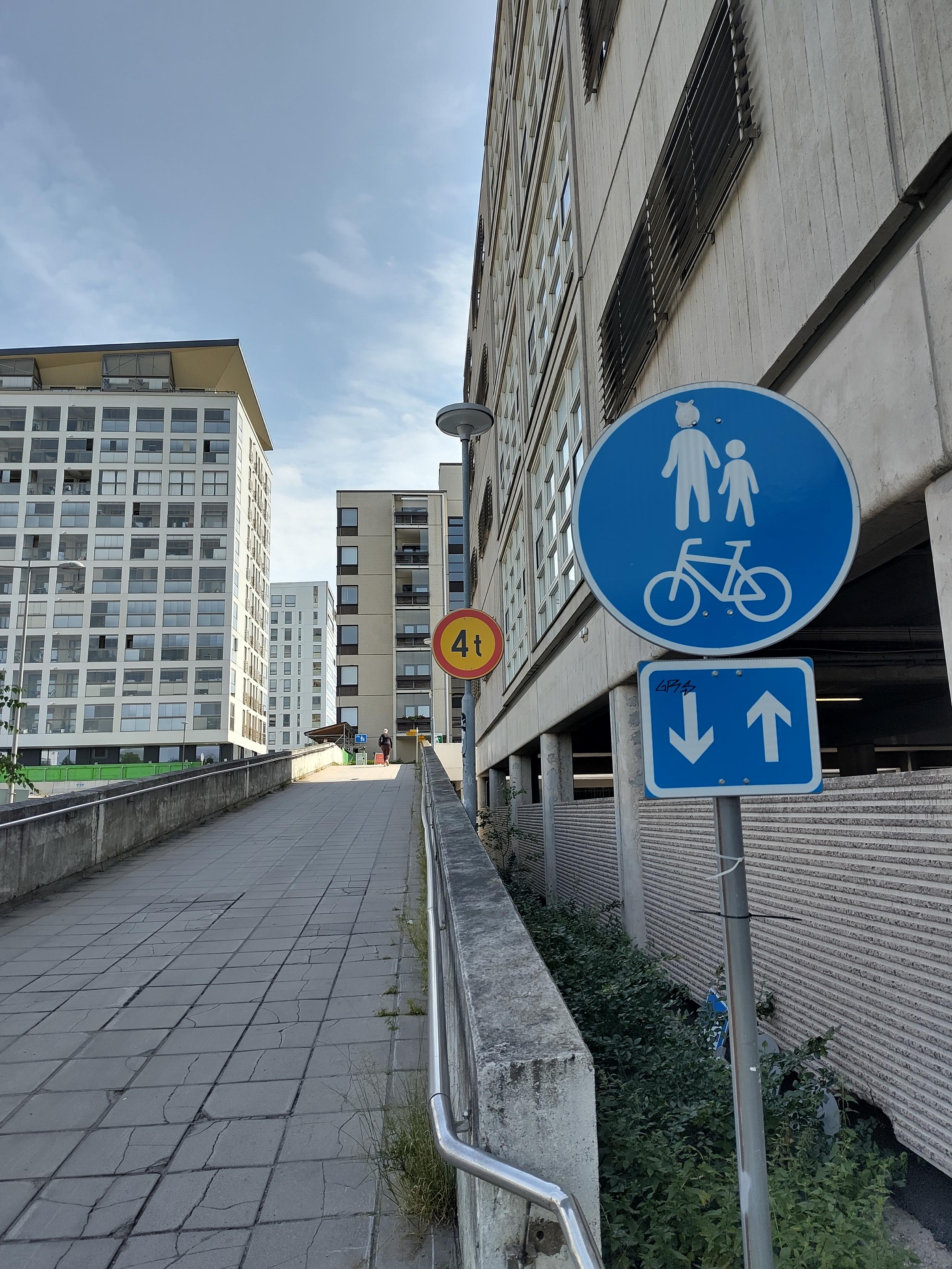 Kuva ylösmenevästä betonirampista kauniina kesäpäivänä. Rampin alussa on liikennemerkki "Yhdistetty jalkakäytävä ja pyörätie" ja sen alla vaikutusalueen merkki merkistä eteen- ja taaksepäin.

Kymmenisen metriä näiden takana on rajoitusmerkki "4 t".

Oikeasta reunasta keskelle menee betonitalon seinä rampin oikealla puolella. Taaempana näkyy asuinkerrostaloja.