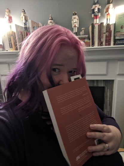 Photo of myself in front of my home fireplace to show off my new hair. Books and nutcrackers are on the mantle. Vibrant pink hair is on top of my head with a middle part; it later fades into a deep purple, styled with some soft curls that go just barely past the shoulder. A book obscures my face from the tip of my nose down. (The book itself is Walking the Clouds, an excellent anthology of indigenous futurist/SF short stories that I've been reading.)