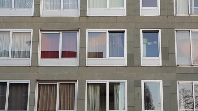 Three rows of white plastic framed, double windows with curtains, the top and bottom row only partially in frame, in the facade of grey concrete stone building. The curtains have a variaty of colors. There are dark and light brown and various shades of green and white ones. Most of them are closed, some half open.