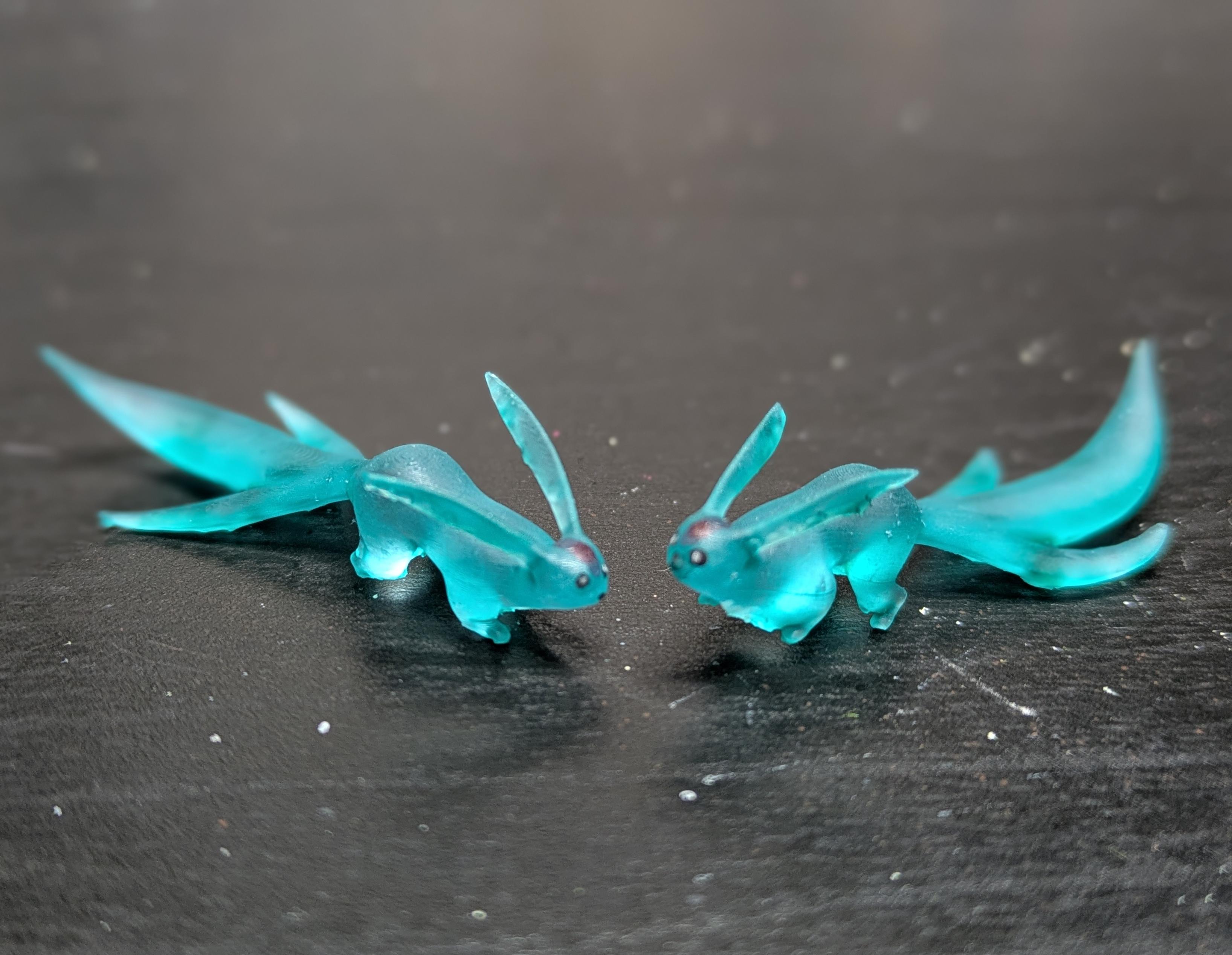 A pair of teal translucent bunny box creatures, with tiny eyes and a red spot on their forehead. They are on a black desk.