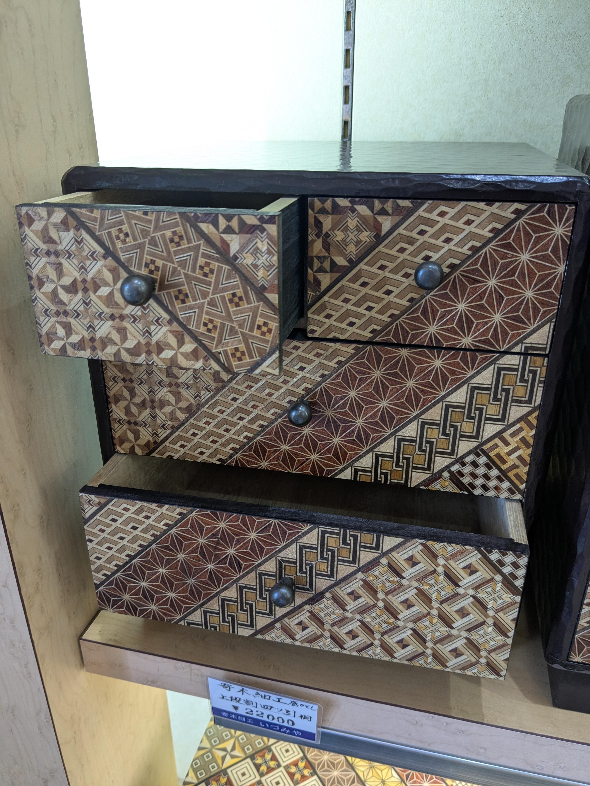 A set of drawers with yosegi designs on the front sides. Three rows of drawers, with two drawers in the top row and one each on the other rows. The top-left and the bottom drawers have been opened slightly.

The yosegi patterns are made from several tiny pieces of wood in different colors to make geometric patterns. There are diagonal stripes across the front of the drawers, with a different geometric design in each stripe. The stripes continue across the gaps between drawers.

A price tag beneath the set reads ¥22,000.