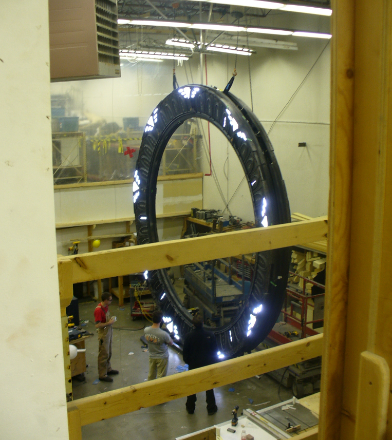 A Stargate being constructed in a warehouse for the TV show Stargate Universe. There are 3 people standing in front of the gate, and the picture is taken from a second floor balcony looking down on them.