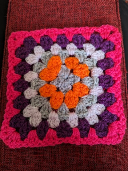 A picture of the blanket so far. 6 rows of granny square crochet. Grey, orange, grey, light pink, purple, and neon pink.