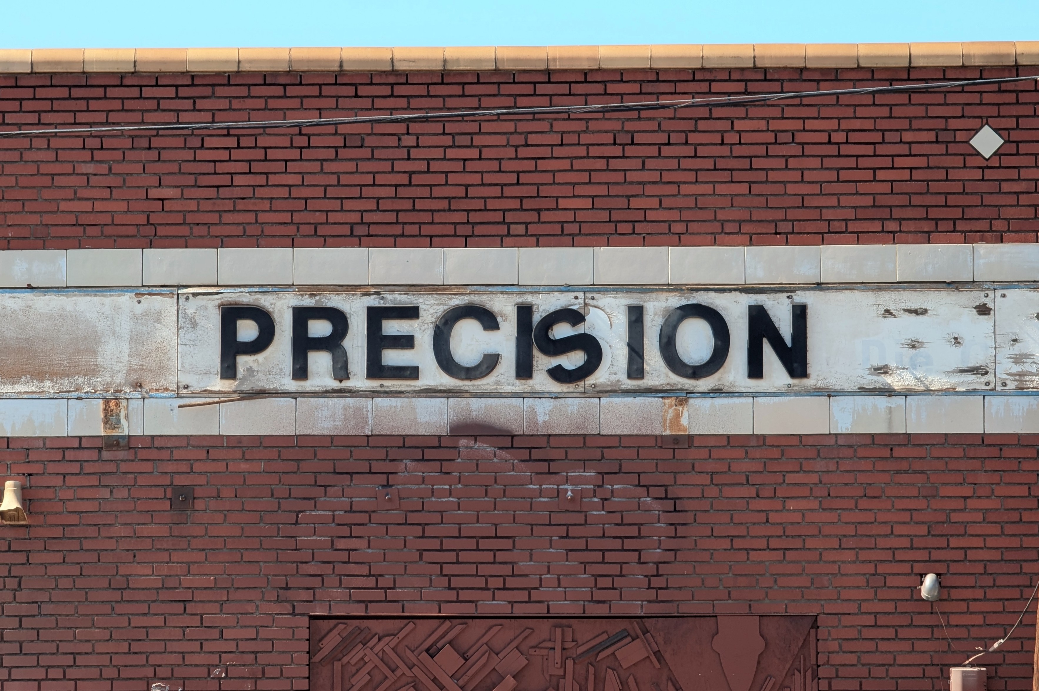 Red brick wall of an old warehouse with a company sign that reads "PRECISION" with the 'S' having fallen comically askew due to age. Graffiti remnants, cables and hardware, and urban found-object art adorn the wall.