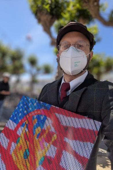 Photo of a person carrying an LED sign, about 0.5m wide, near some trees. The sign is displaying a flag and some emojis.