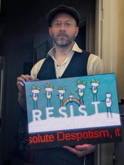 a man wearing a flat cap, button up shirt and vest, holding a LED signboard featuring dancing frogs