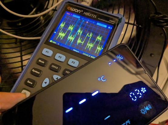 Picture of an oscilloscope multimeter showing a very noisy PWM signal. behind is the cage of a fan, in front is the fan control panel