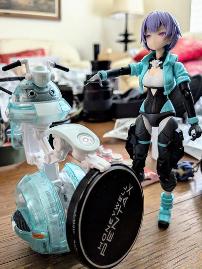 A light teal colored robot is holding a Pentax lens cap. A purple haired anime-styled girl is standing next to it, pointing towards the robot.