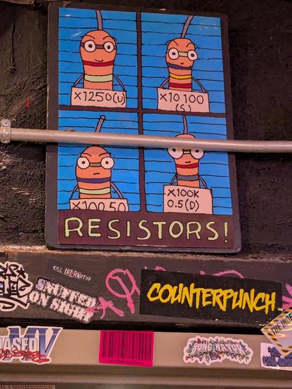 A poster board held in place with electrical conduit on the sticker-filled wall. The poster shows 4 electrical resistors in the style of police mug shots. Below, the poster is captioned "Resistors!"