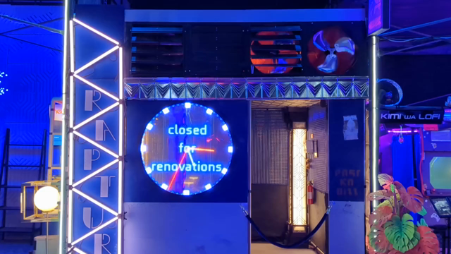 What looks like a small building basked in blue light. There are letters "RAPTURE" inside a set of glowing triangles to the left. In the middle is an LED analog clockface. Above is a set of 4 exhaust fans, two are spinning slowly. Inside a glowing column is visible in the back corner. The entry is closed off with a velvet rope.