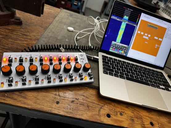 A close-up shot shows a music production setup with a hardware controller and a laptop on a wooden workbench. The white hardware controller on the left has eight vertical columns labeled "out 1", "out 2", "out 3", "out 4", 4", "out 5", "out 6", "out 7", and "out 8" at the top, and numbered "1" through "8" at the bottom, each featuring an orange knob and lights. To the right, a laptop displays two software windows: one with a blue and yellow waveform and another with an orange interface. White cables connect the devices across the worn wooden surface. The background shows blurry workshop tools and equipment.