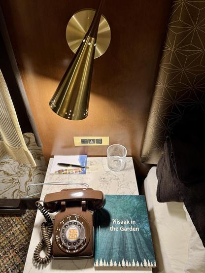 A brown dial telephone on a hotel bedside table, surrounded by other midcentury items: a wall lamp, the drapes, and the headboard.