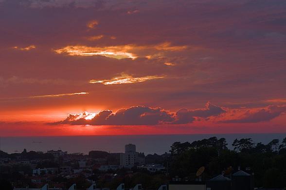 Red sky, sun and clouds in the background above a dark ocean with dark buildings lower down in the foreground.