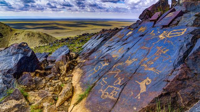A close view of petroglyphs on sloping rocks overlooking the Gobi Desert grasslands from a high ridge. The petroglyphs portray animals and other figures.