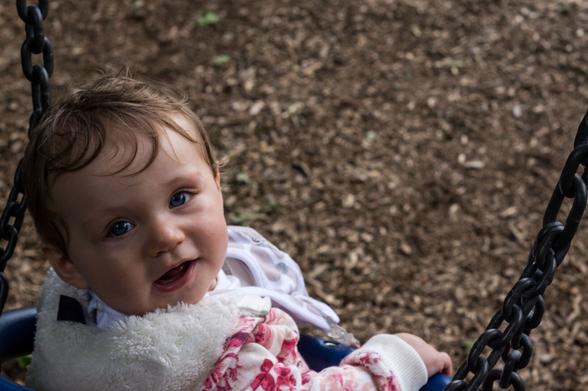 A young girl with a white and pink jumper looks up at the camera, in the background is woodchip ground.  The chain of the swing crosses the bottom right corner.