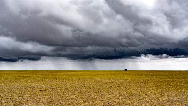A distant view of massive gray rain clouds behind a small hut near the horizon. The foreground is short matted yellow-green grasses extending to the horizon at lower mid-frame.