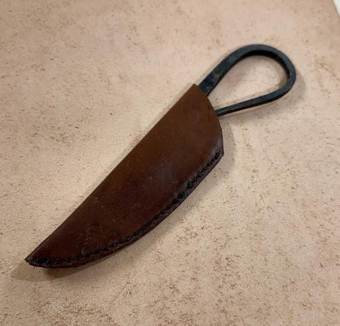 A hand forged knife with a black handle partially encased in a brown leather sheath on a beige surface.