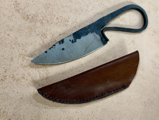 A hand forged curved knife with a ring handle beside a brown leather sheath on a textured surface.