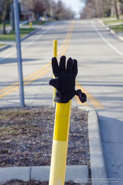 A black winter glove appears to be offering a high-five from the top of bright yellow post that has been inserted into it. In the background are concrete curbs and a signpost, and another yellow reflective post. In the distance, yellow and white lane stripes extend past a yellow fire hydrant, up a hill into the distance.
