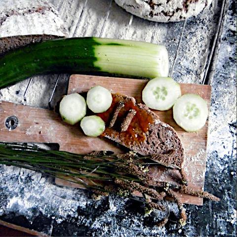 Das Bild zeigt einen bemehlten Tisch mit einem Schneidebrett, darauf liegt eine Scheibe Heubrot mit Mirabellen,armelade und mit Gurke belegt, Spitzwegerichblüten dazu.