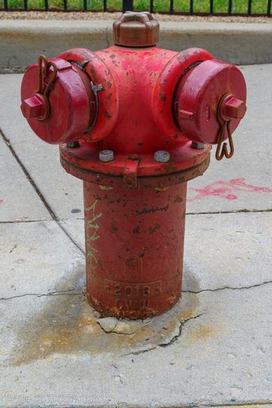 A red fire hydrant with two ports and a brass security nut on top emerges from the cracked pavement of a sidewalk. the paint is a little chipped and weathered, and rust can be seen in areas where the paint is missing. The base of the hydrant is embossed with  a globe logo with a lower-case e  beneath it, the year "2016," and the letters "CWW." Some green grass can be seen through a wrought-iron fence in the background.